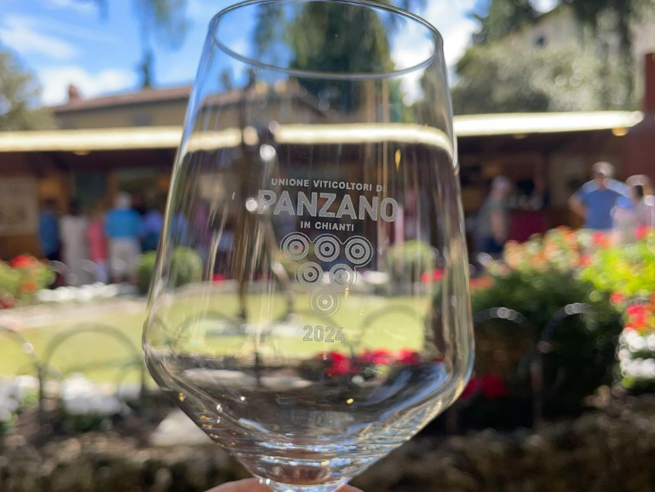 Un calice da vino in vetro trasparente tenuto in primo piano, con inciso il logo "Unione Viticoltori di Panzano in Chianti" e l'anno "2024". Sullo sfondo, leggermente sfocato dal gioco di messa a fuoco, si intravede un giardino all'aperto durante un evento con persone e stand espositivi sotto il sole.