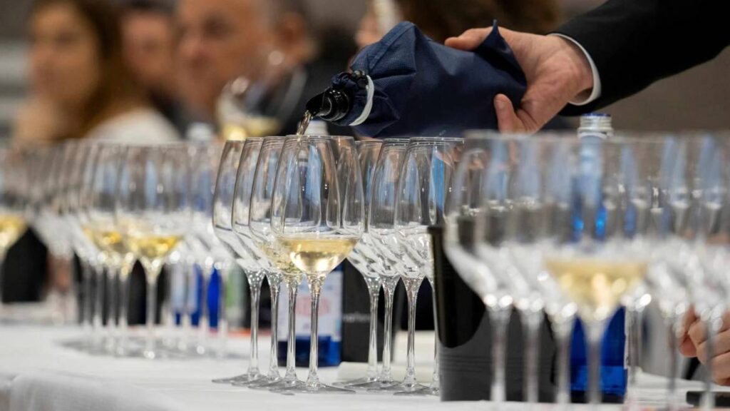 Sommelier che versa vino bianco in una serie di calici allineati su una tavola apparecchiata, durante una degustazione guidata con partecipanti seduti sullo sfondo.