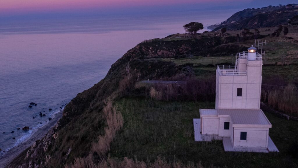 Faro sul promontorio Rasocolmo una scogliera erbosa affacciata sul mare al tramonto, con cielo viola‑rosato e costa rocciosa sottostante.