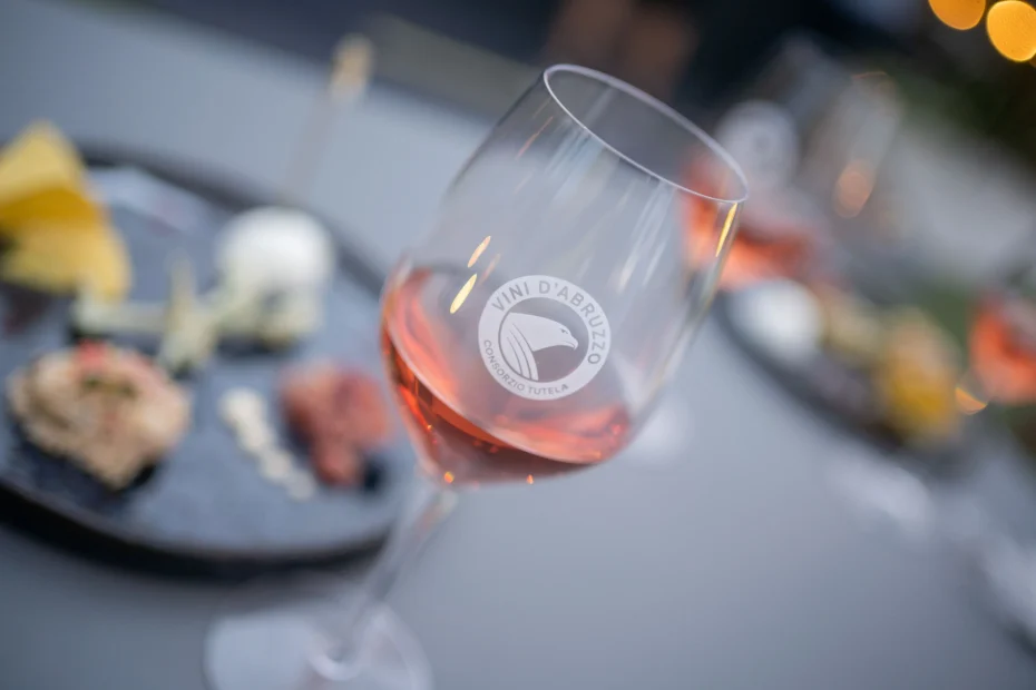 Cerasuolo d'Abruzzo a Roma, il Wine Experience a Ripetta Palace 18 giugno 2026. Primo piano di un calice di vino rosato con il logo “Vini d’Abruzzo – Consorzio Tutela”, nitido contro uno sfondo sfocato con piatti di salumi, formaggi e pane, evocando una scena di degustazione enogastronomica.