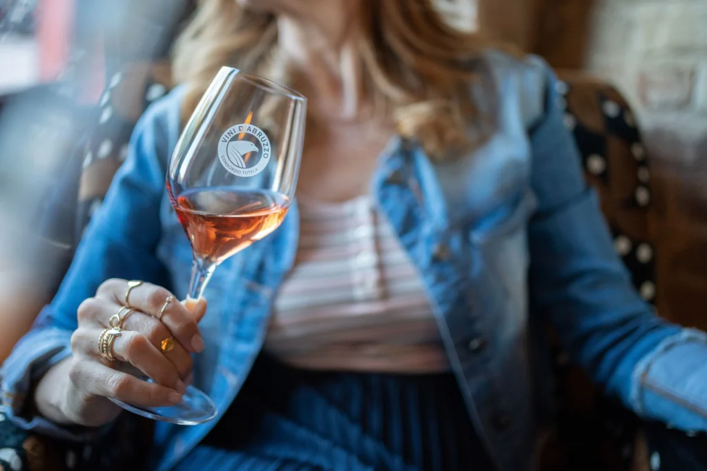 Cerasuolo d'Abruzzo a Roma, il Wine Experience a Ripetta Palace 18 giugno 2026. Immagine di una donna che tiene in mano un calice di vino rosato con il logo “Vini d’Abruzzo – Consorzio Tutela”. La persona indossa una giacca di jeans sopra una maglia a righe; lo sfondo è caldo e sfocato, suggerendo un ambiente accogliente da degustazione.