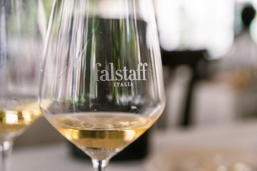 Falstaff White Wine Party Roma. un calice da degustazione con vino bianco dal colore dorato, marchiato falstaff Italkia in primo piano. Sullo sfondo sfocato si intravede un secondo calice e un ambiente da degustazione o ristorante