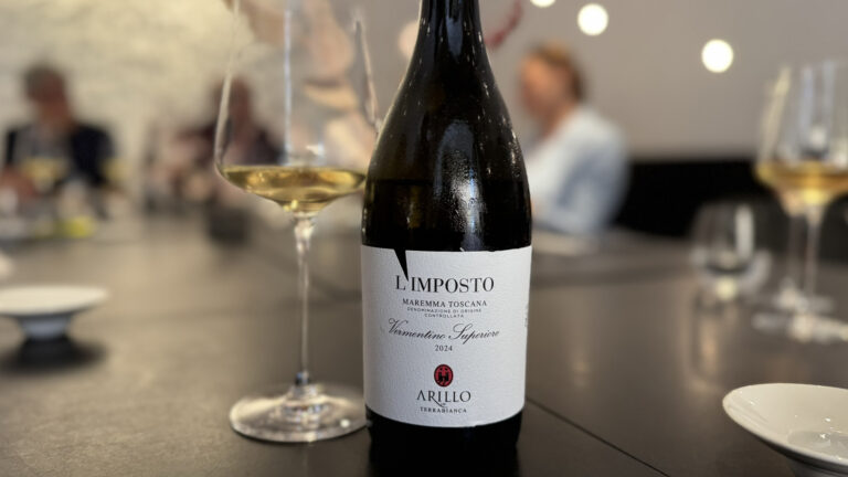 Bottiglia di Vermentino Superiore 2024 L’Imposto di Arillo in Terrabianca in primo piano accanto a un calice di vino bianco sul tavolo del ristorante Per Me di Giulio Terrinoni durante il Press Lunch organizzato a Roma. Sullo sfondo sfocato si intravedono persone sedute, creando un’atmosfera da evento enologico professionale.