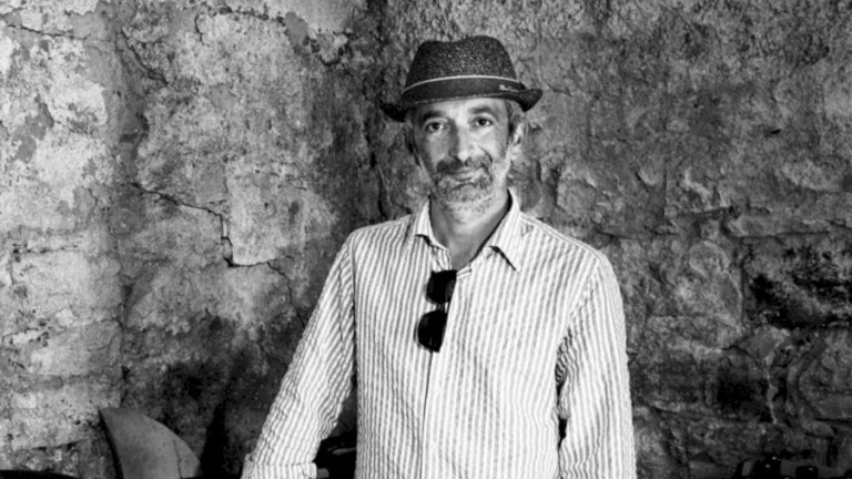 Luigi Tecca, produttore di vino naturale del movimento Triple A, in piedi davanti all'interno della sua cantina. Vi vedono un muro di pietra in un ambiente rustico con cassette contenenti bottiglie di vino ed una panca di legno contenente anfore di terracotta.