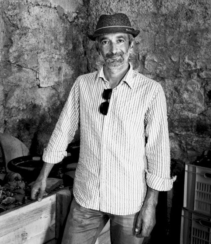 Luigi Tecca, produttore di vino naturale del movimento Triple A, in piedi davanti all'interno della sua cantina. Vi vedono un muro di pietra in un ambiente rustico con cassette contenenti bottiglie di vino ed una panca di legno contenente anfore di terracotta.
