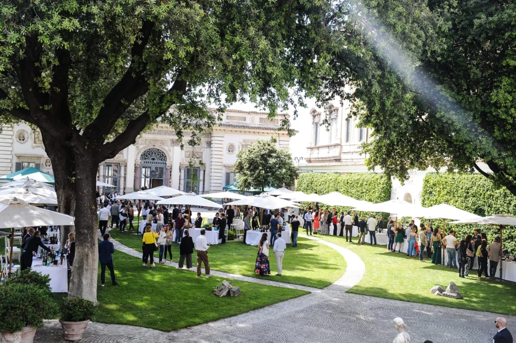 Vino e Arte che Passione al Casino dell'Aurora Pallavicini. Ampia veduta del giardino storico del Casino dell'Aurora Pallavicini a Roma durante l'evento "Vino e Arte che passione". Numerosi partecipanti passeggiano e si intrattengono sul prato verde e lungo un camminamento in pietra curvo. L'area all'aperto è allestita con grandi ombrelloni bianchi che riparano i banchi d'assaggio ricchi di bottiglie di vino per le degustazioni. Sullo sfondo si erge la facciata barocca del Casino dell'Aurora, arricchita da rilievi classici e ampie vetrate, parzialmente ombreggiata dalle fronde di grandi alberi secolari.